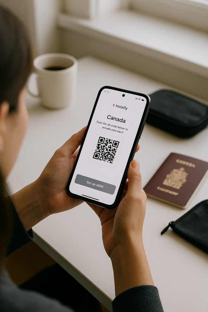 Over-the-shoulder view of a person holding a smartphone showing the Holafly eSIM Canada activation screen. A QR code is visible on the phone, with a Canadian passport, coffee mug, and travel pouch on a white desk under soft daylight.